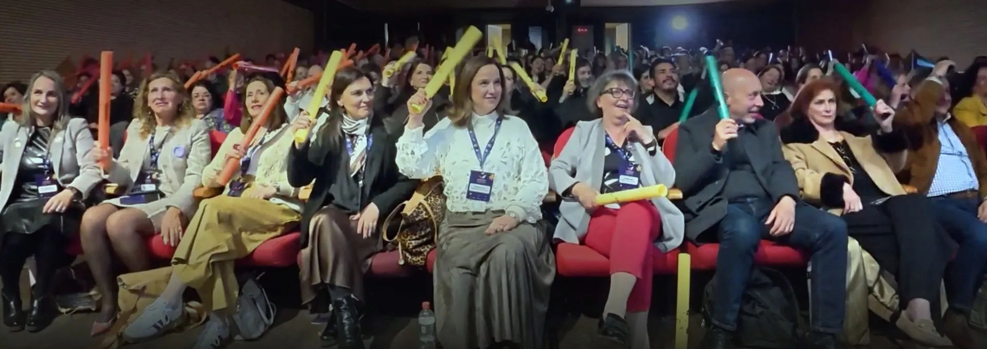 Público sonriente en un auditorio, levantando tubos de colores durante una experiencia participativa.