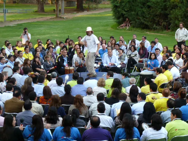 Un facilitador de sombrero blanco, de pie al centro, dirige a un gran círculo de personas sentadas al aire libre que tocan tambores y percusión.